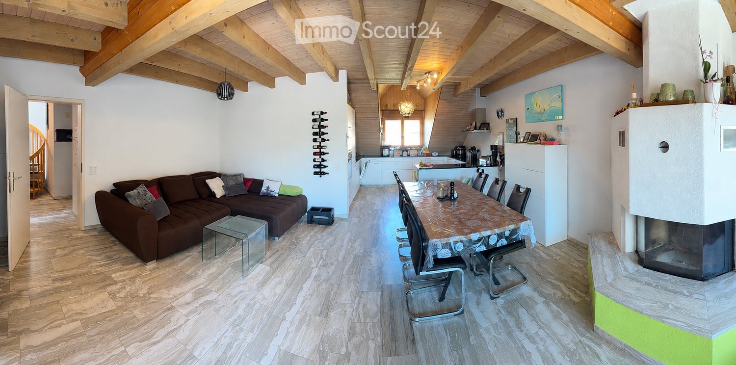 Open-plan living and dining area with exposed wooden beams, a brown sectional sofa, a glass coffee table, a dining table with chairs, and a modern fireplace. The kitchen area features white cabinets and stainless steel appliances.