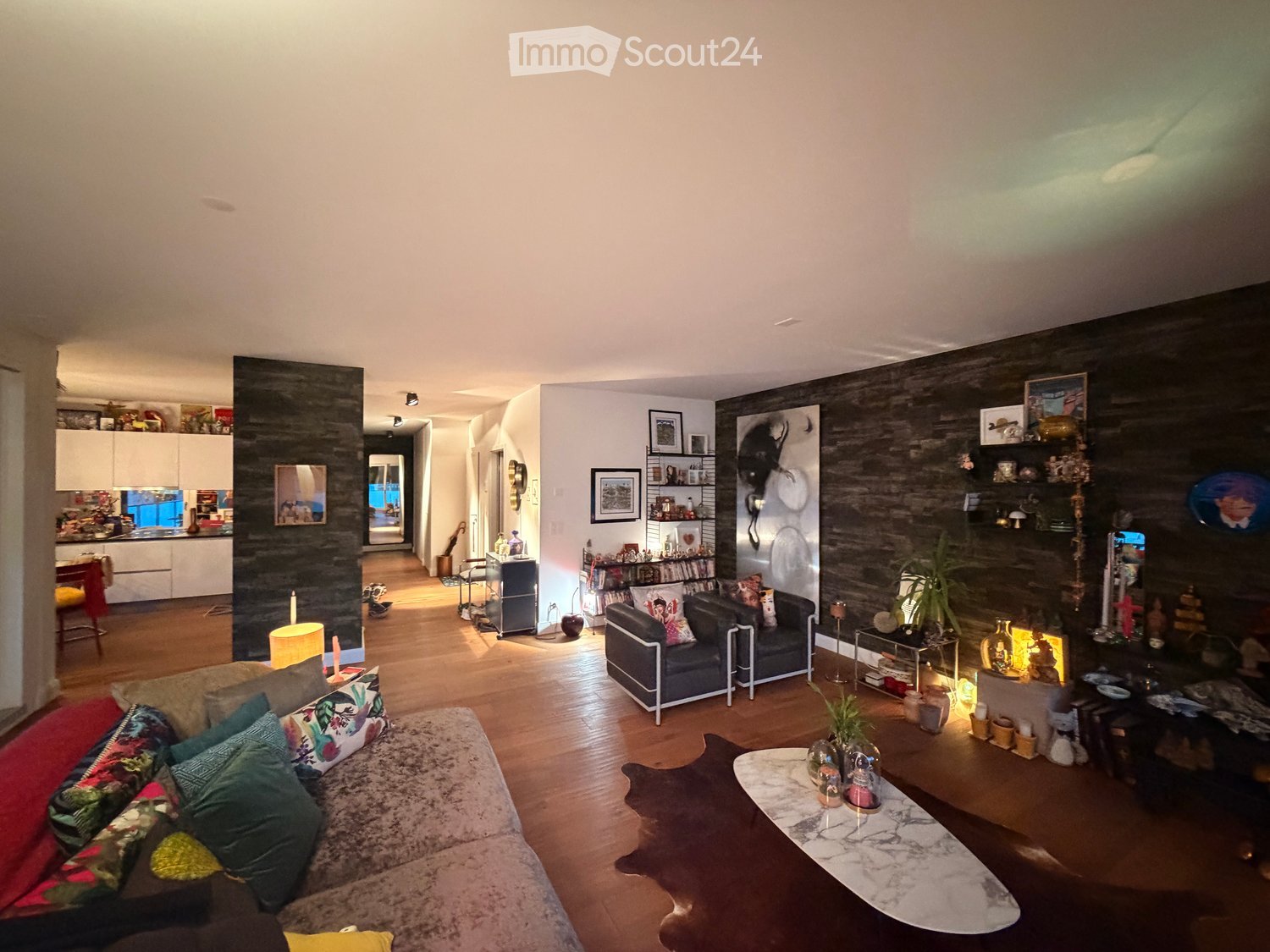 Living room with wooden flooring, a large sofa with colorful cushions, a coffee table, a side table, shelves with decorative items, and framed pictures on the wall.