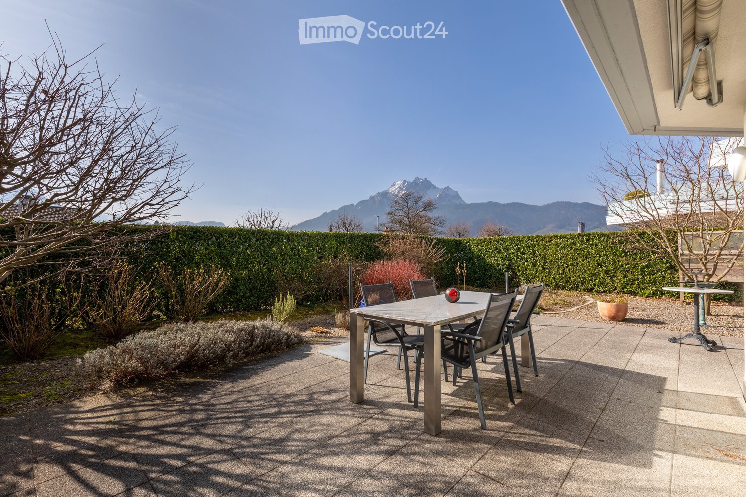 Outdoor patio with a table and chairs, surrounded by a hedge and a view of mountains.