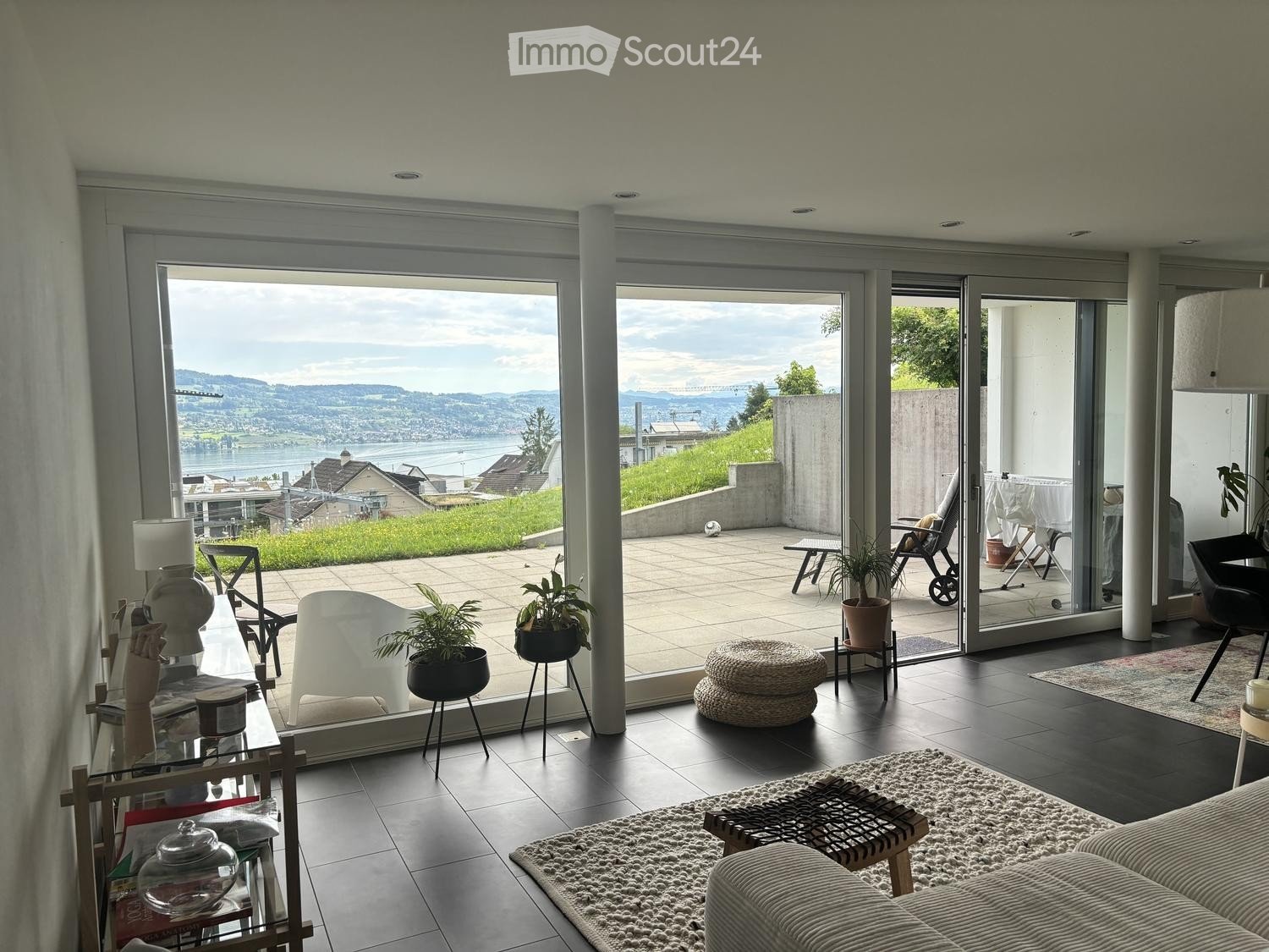 Modern living room with large glass doors opening to a balcony, offering a scenic view of a lake and hills. The room features a sofa, coffee table, chairs, a lamp, and potted plants.