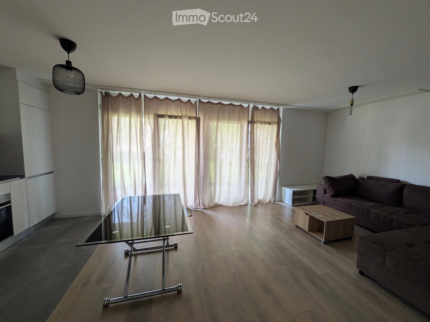 Modern living room with wooden flooring, large windows covered by beige curtains, a glass coffee table, a dark brown sofa, and a small wooden side table.