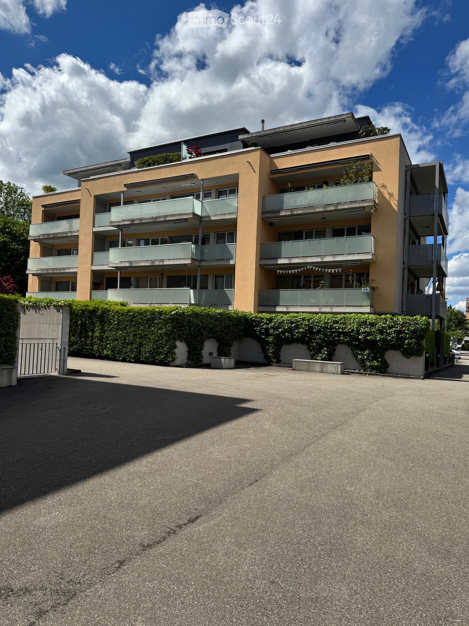 Modern multi-story residential building with balconies, surrounded by greenery and a paved driveway.