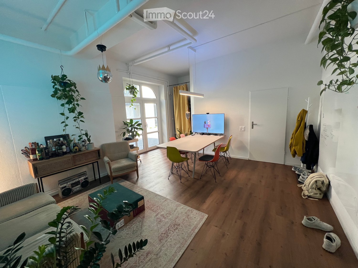 A modern, well-lit living room with wooden flooring, a large window, and a mix of seating and dining areas. Features include a sofa, armchair, coffee table, dining table with chairs, a television, and several plants.