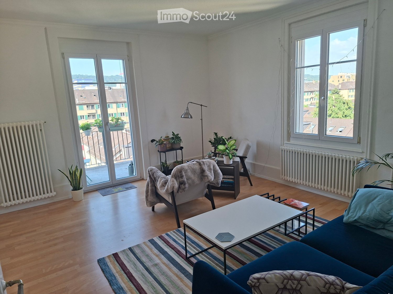 Bright living room with wooden flooring, large windows, and a balcony. Features a blue sofa, white coffee table, armchair with a fur throw, floor lamp, and several potted plants.