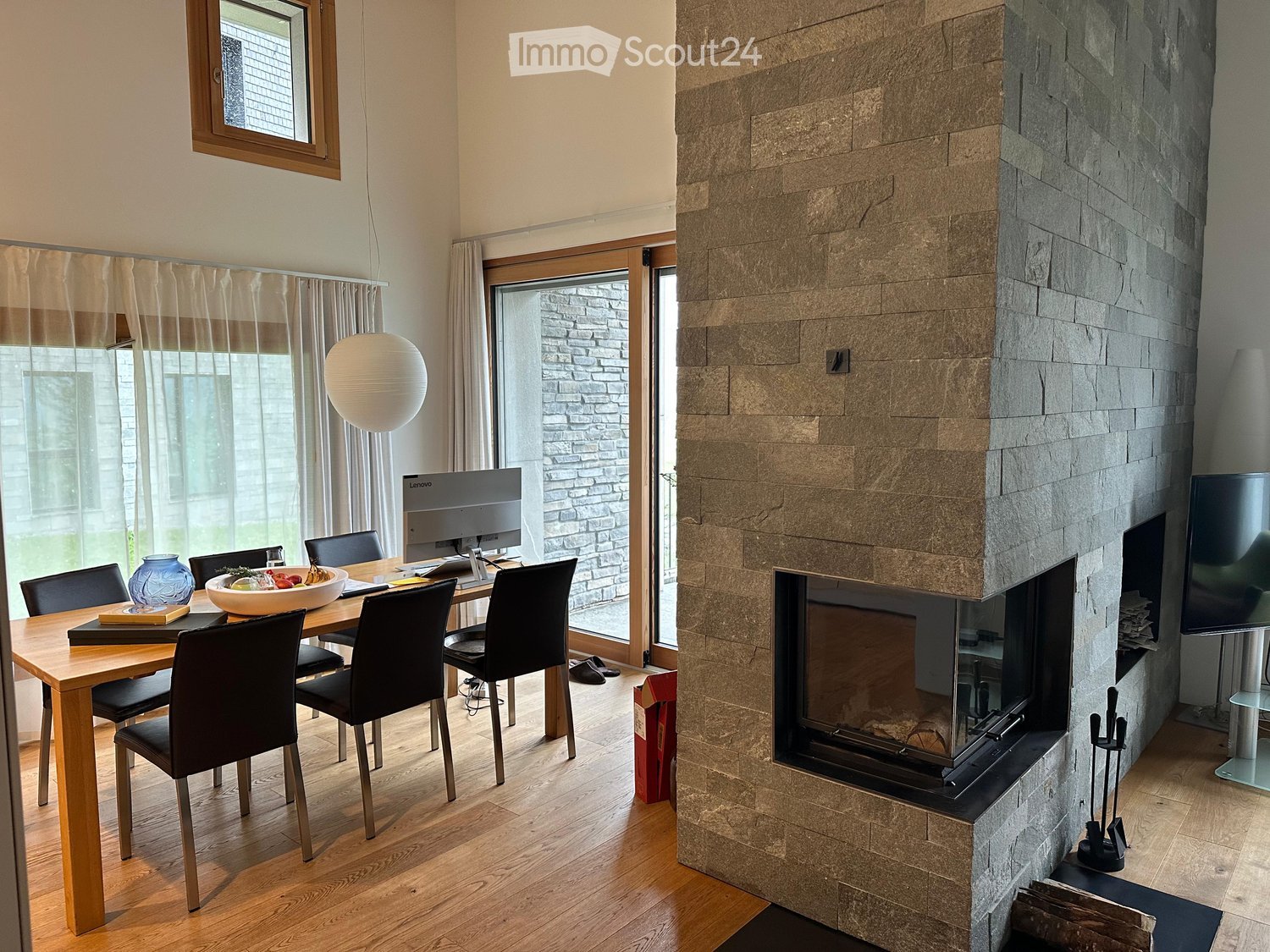 Modern living and dining area with a stone fireplace, wooden dining table, chairs, and a large screen. Natural light from large windows and a hanging ball lamp illuminate the space.