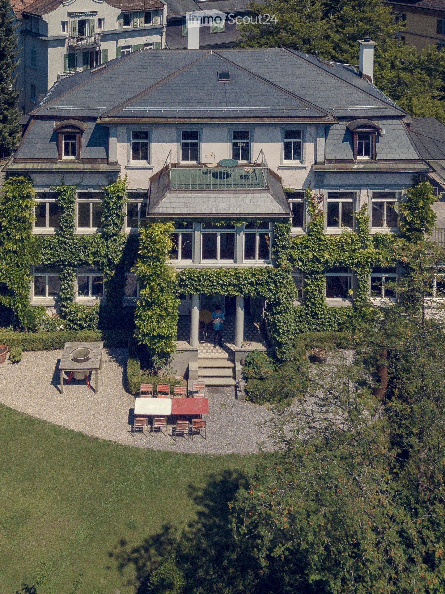 A multi-story house with a grey roof, white walls, and multiple windows. The house features a balcony with outdoor furniture and a well-maintained garden area with a patio and seating arrangement.