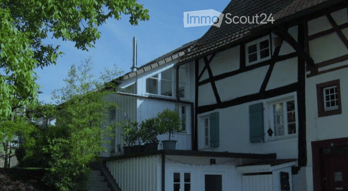 Traditional half-timbered house with white walls, dark wooden beams, multiple windows, and a sloped roof. A small garden with potted plants is visible in front.
