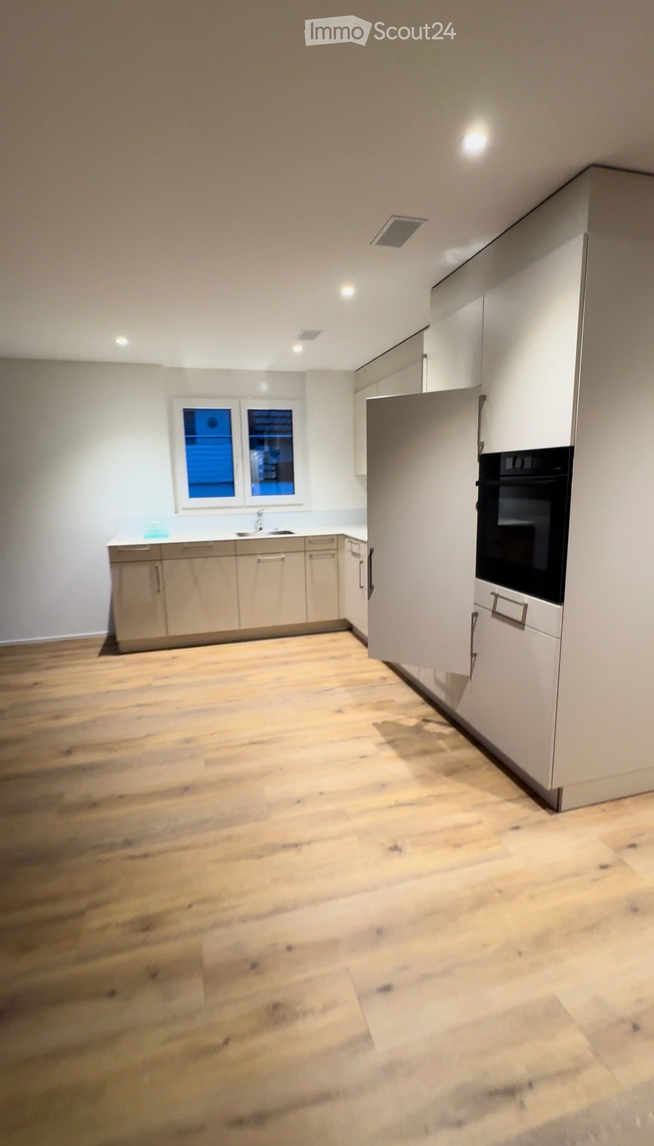 Empty kitchen with light wooden flooring, white cabinets, and a sink area. No appliances or furniture are present.