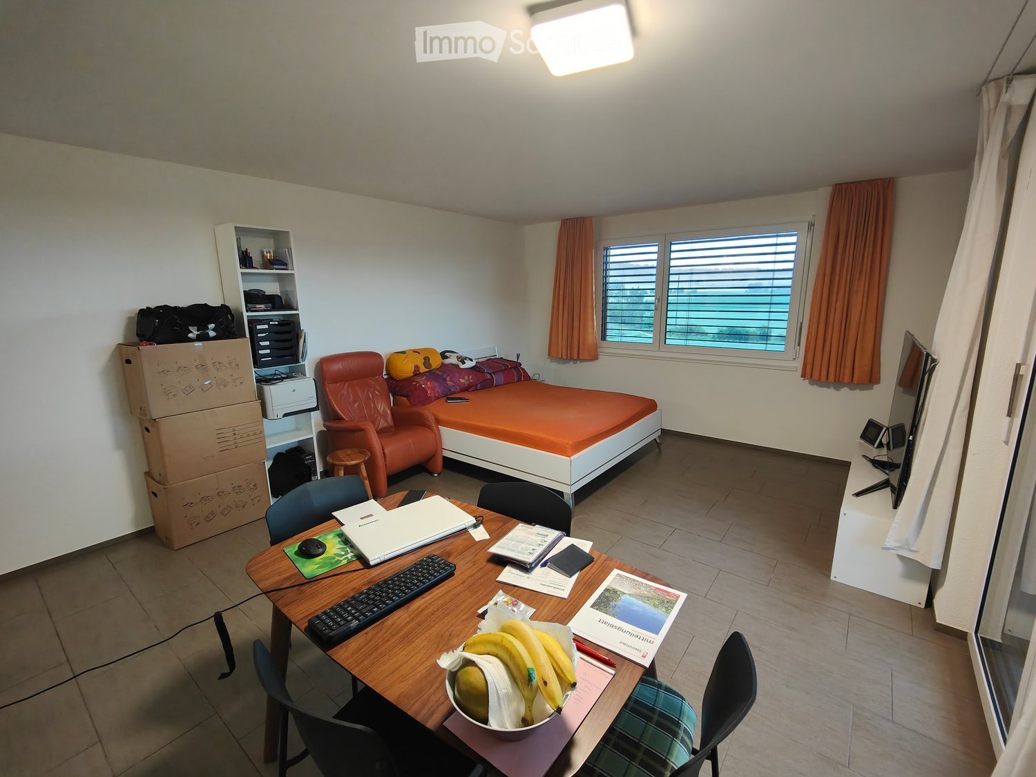 The room is furnished with a bed, a chair, a table with a laptop, keyboard, and mouse, and a screen. There are also curtains, a stool, and a shelf with items on it.