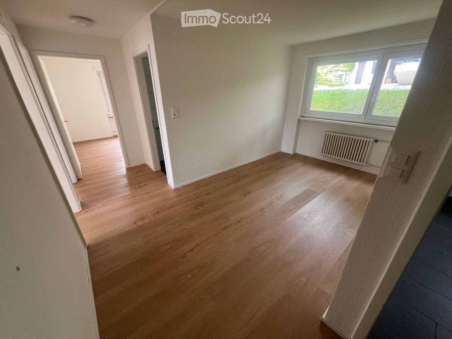 Empty hallway with wooden flooring, white walls, and a window showing a view of greenery outside. No furniture or equipment is present.