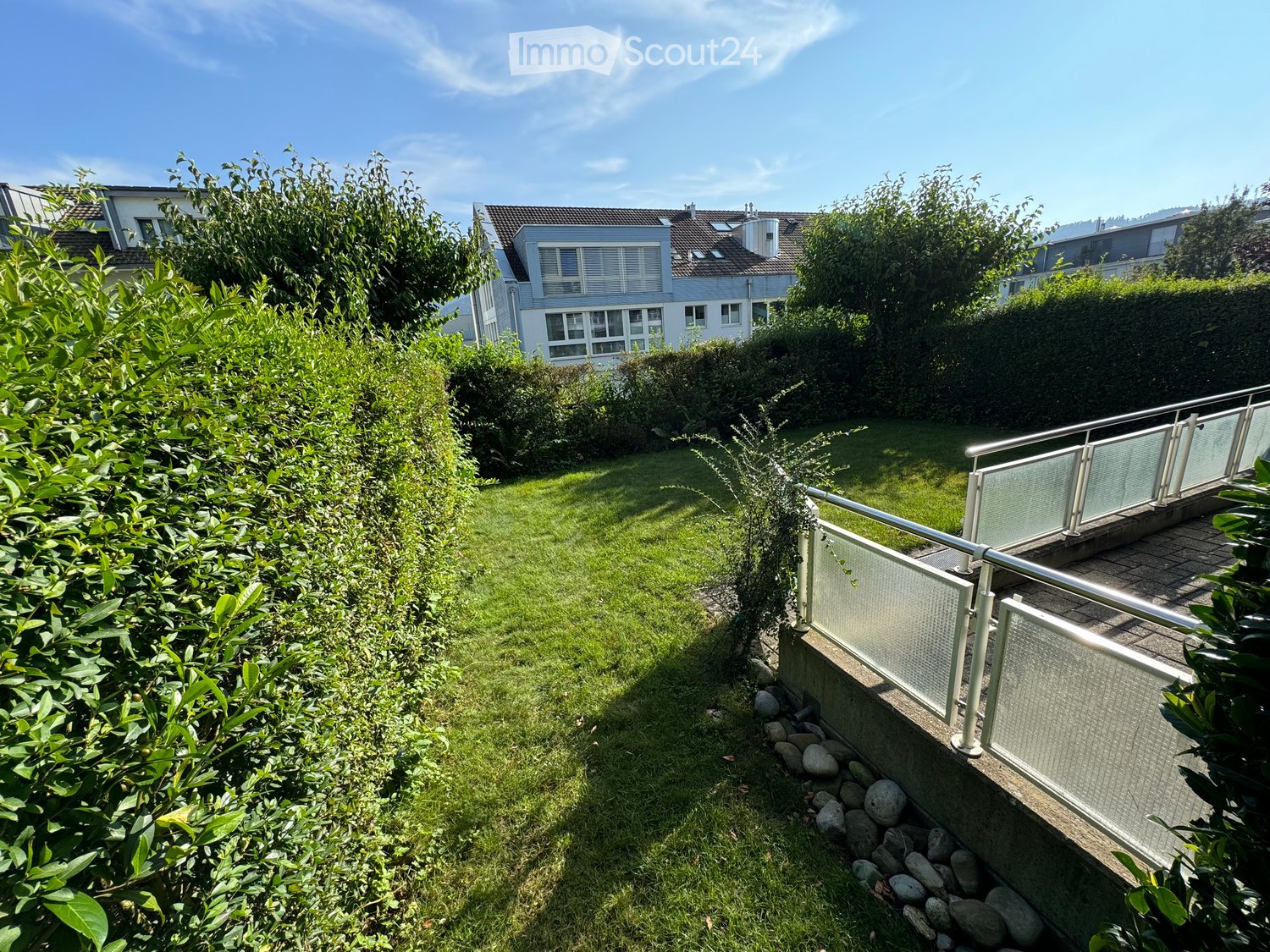 Well-maintained garden with a raised ground floor, surrounded by greenery and a modern railing leading to a patio area.
