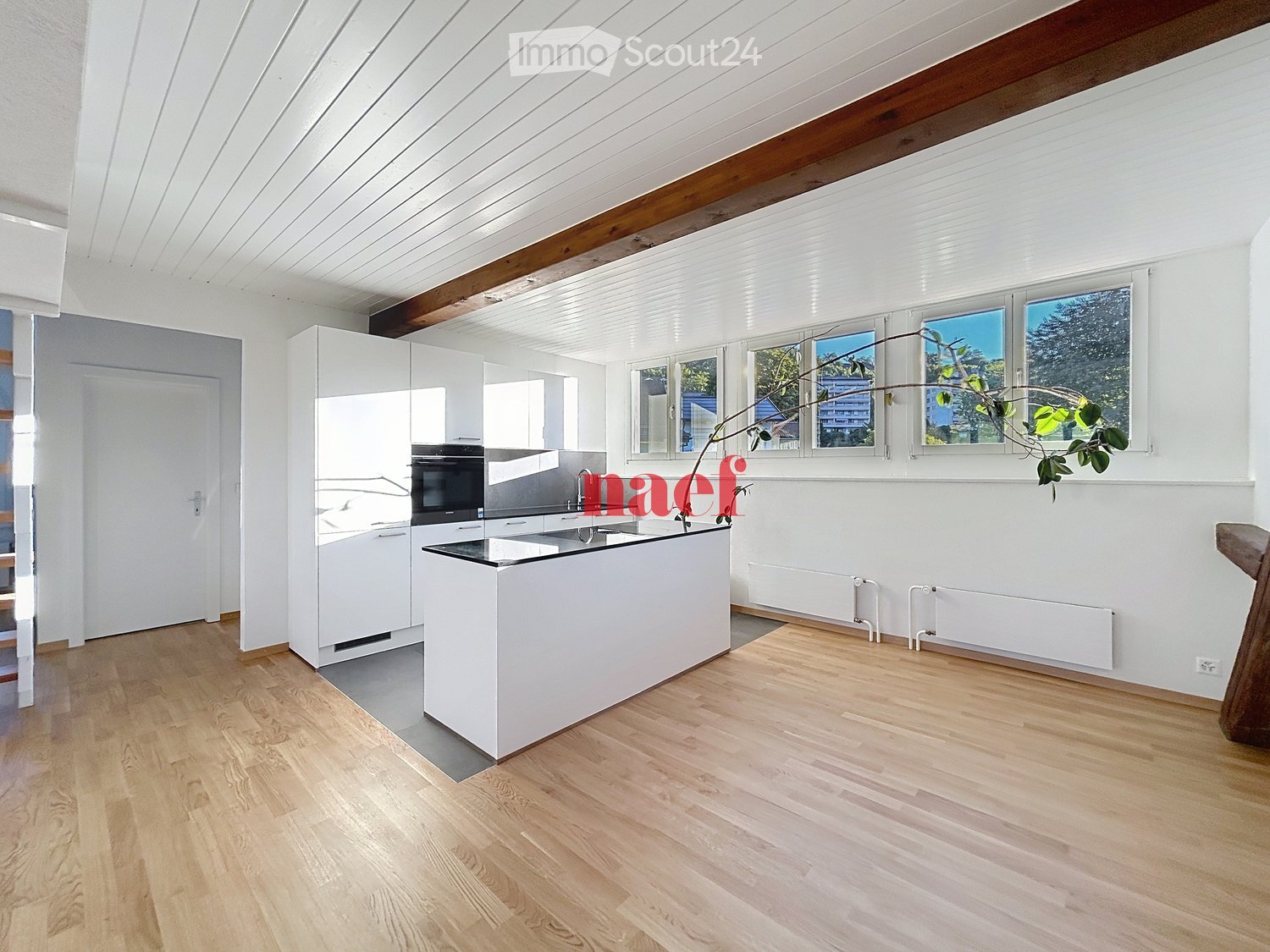 Modern kitchen with white cabinets, a central island, and wooden flooring. Features include an oven, sink, and large windows with a view of greenery.