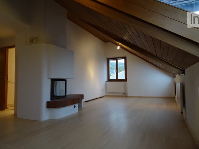 Empty attic room with wooden floors, slanted wooden ceiling, fireplace, and large window with view.