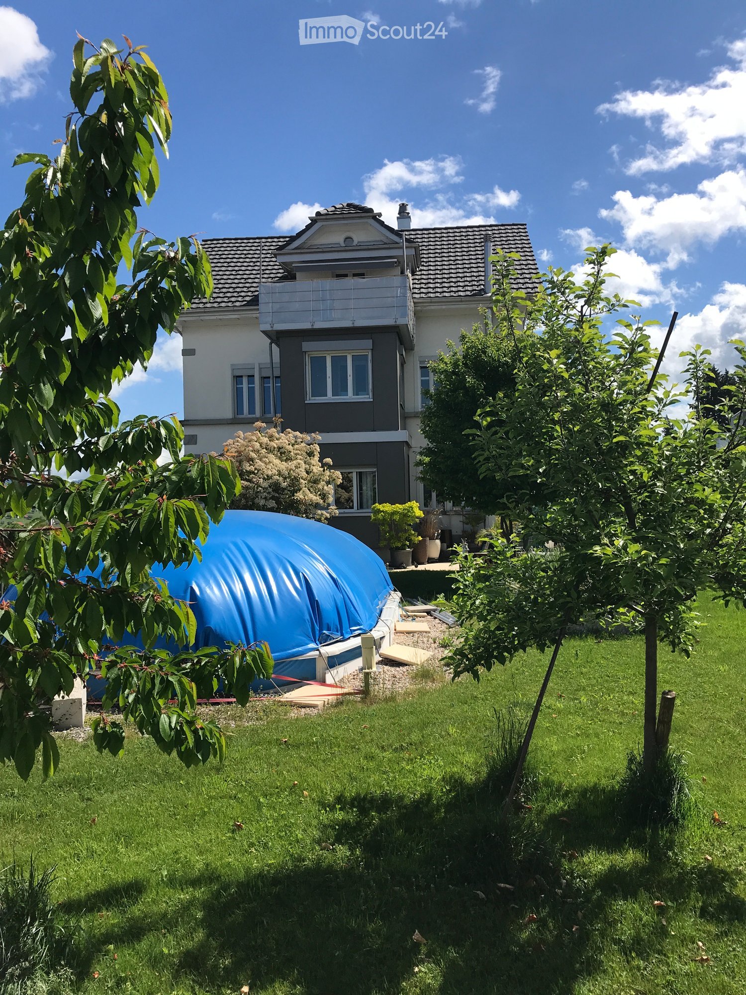 A two-story house with a balcony, surrounded by a well-maintained lawn, trees, and a blue inflatable pool covered with a tarp.