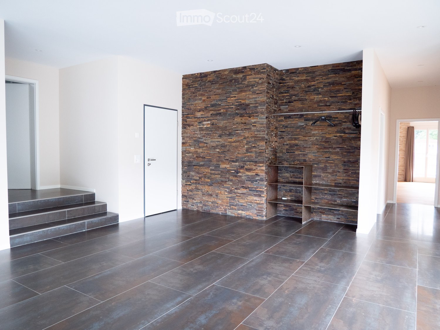 Modern hallway with dark tiled flooring, a stone accent wall, built-in wooden shelving, and a coat rack with hangers.