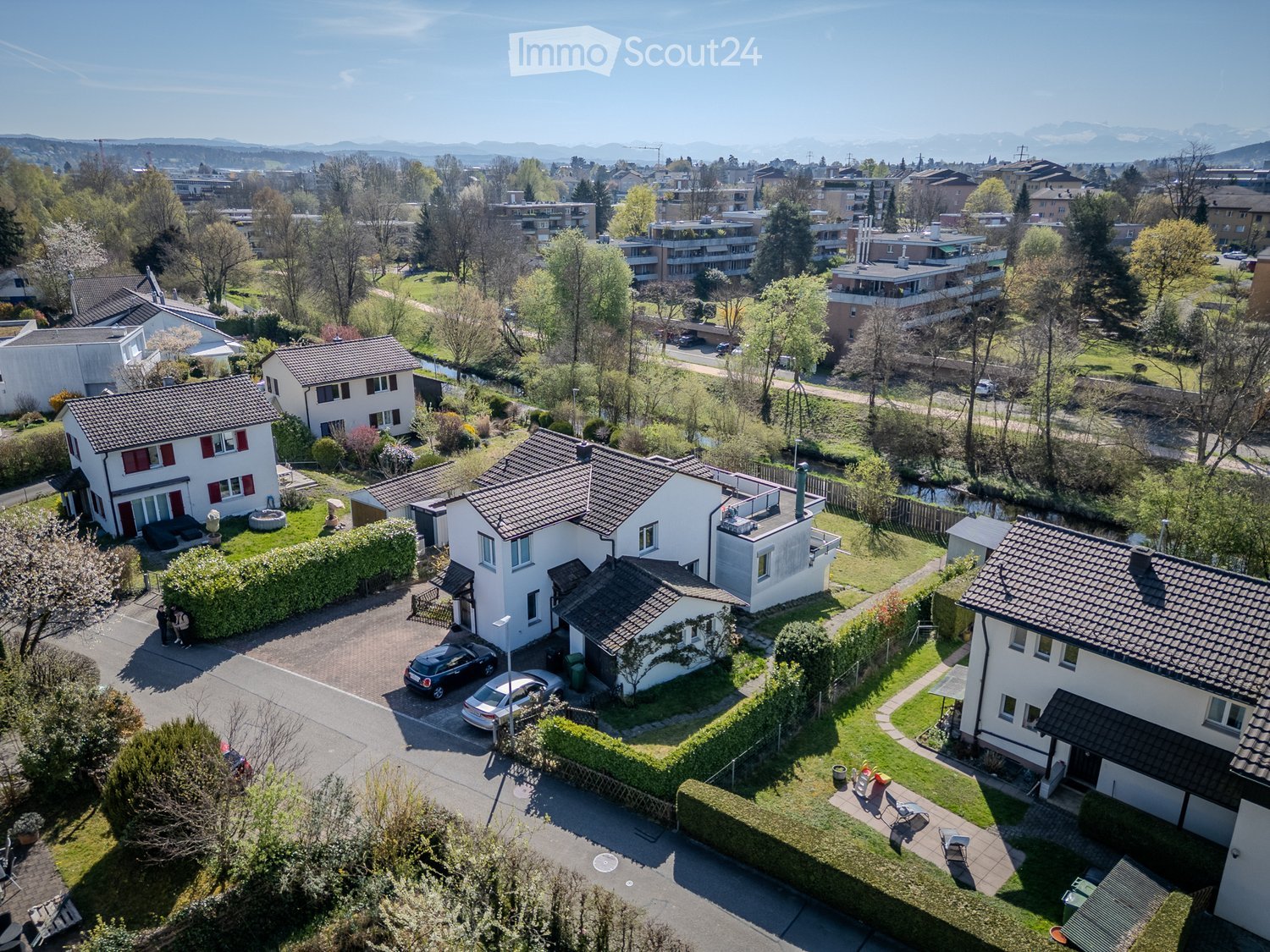 Aerial view of a residential neighborhood with multiple houses, gardens, and parked cars. The houses have tiled roofs and are surrounded by greenery and trees. The area includes a mix of detached and semi-detached homes, with well-maintained lawns and dri
