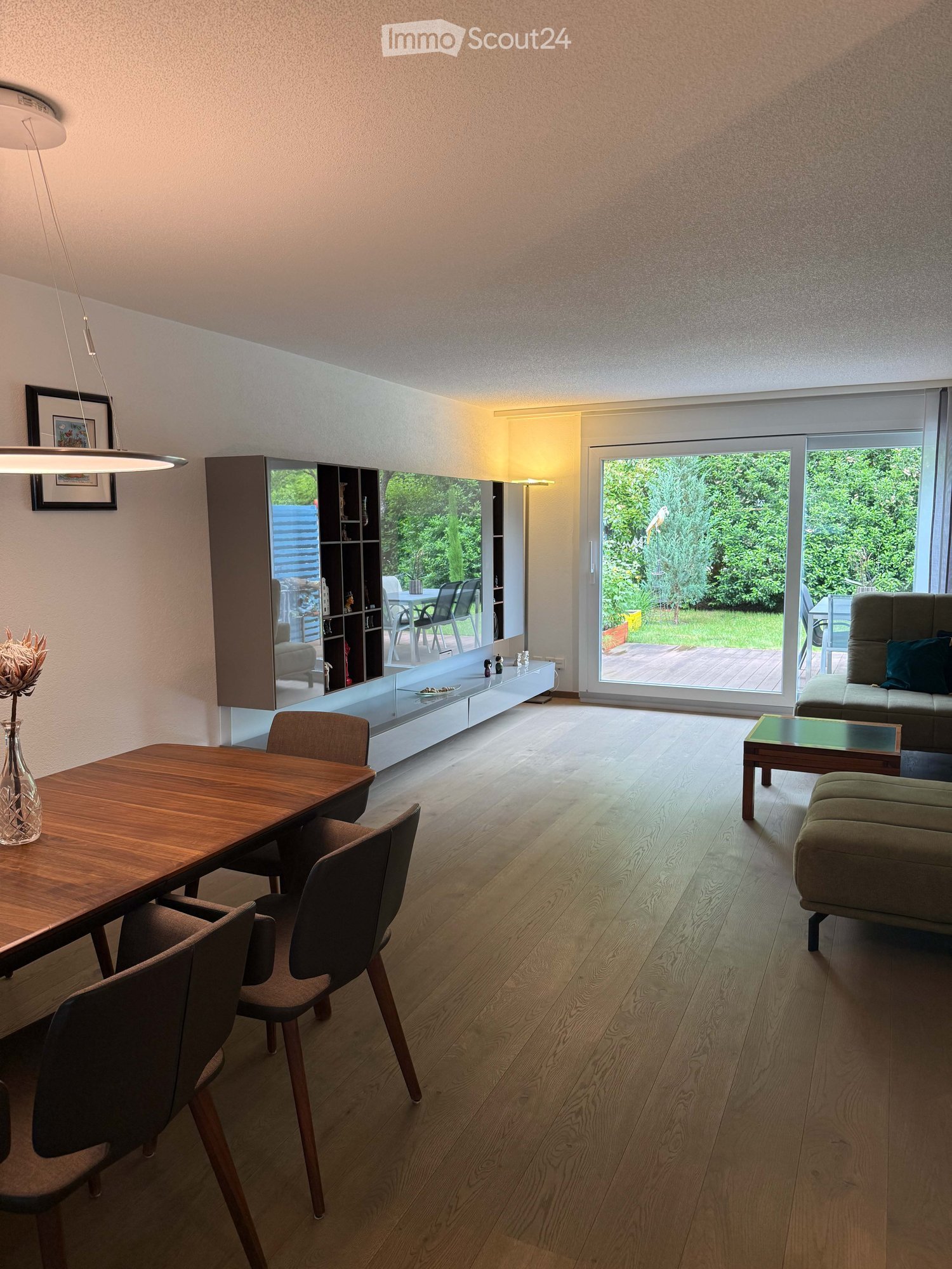 Modern living and dining area with light wood flooring, large windows, and a sliding glass door leading to a garden. Features a sofa, coffee table, ottoman, dining table with chairs, a shelf unit, and decorative vase.