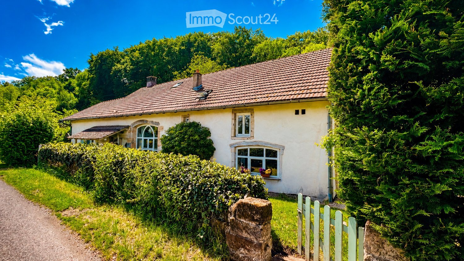 A charming single-story house with a tiled roof, white exterior, and multiple windows, surrounded by lush greenery and a gravel pathway.