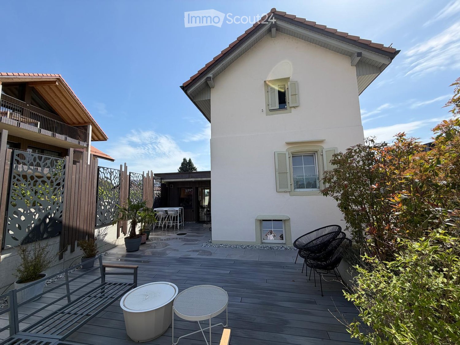 A modern outdoor patio area with wooden decking, a round table, chairs, a bench, and potted plants, adjacent to a two-story house with white walls and shutters.