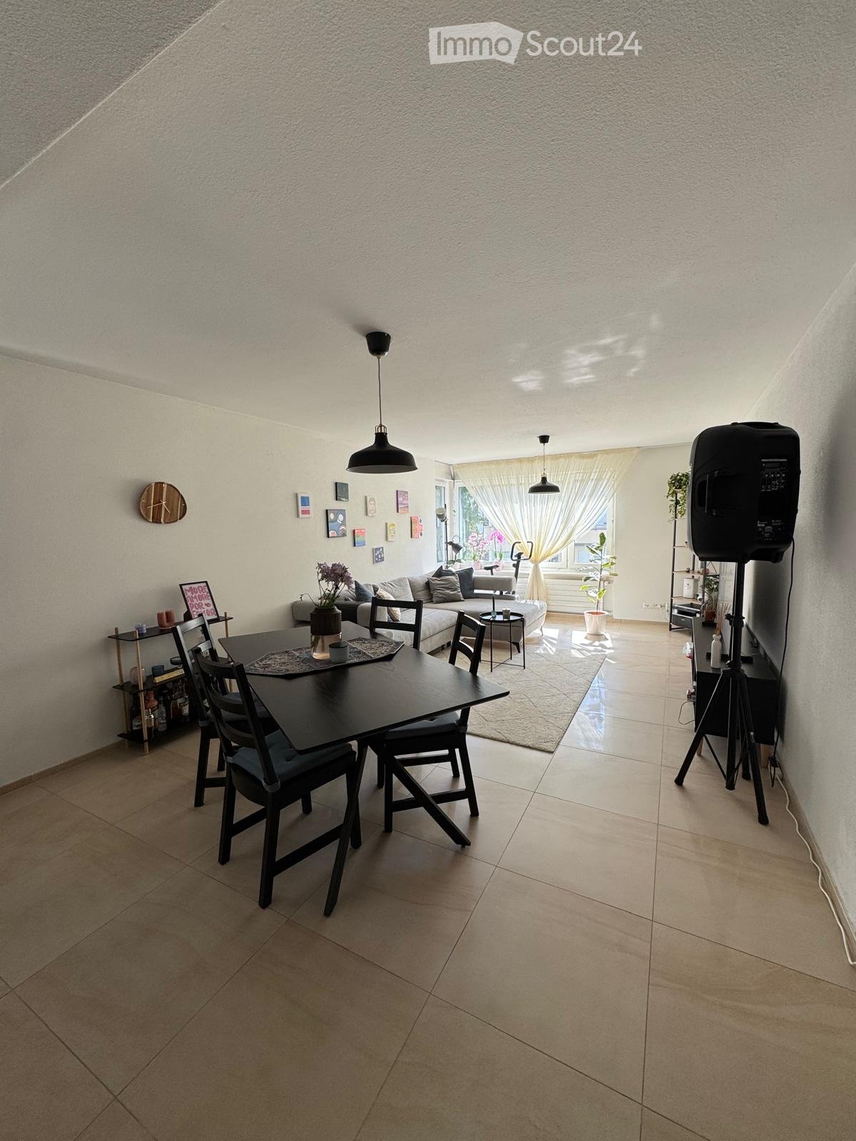 A modern living and dining area with a sofa, dining table, chairs, and a large window with curtains. The space features a tiled floor, pendant lights, and decorative items on the walls.