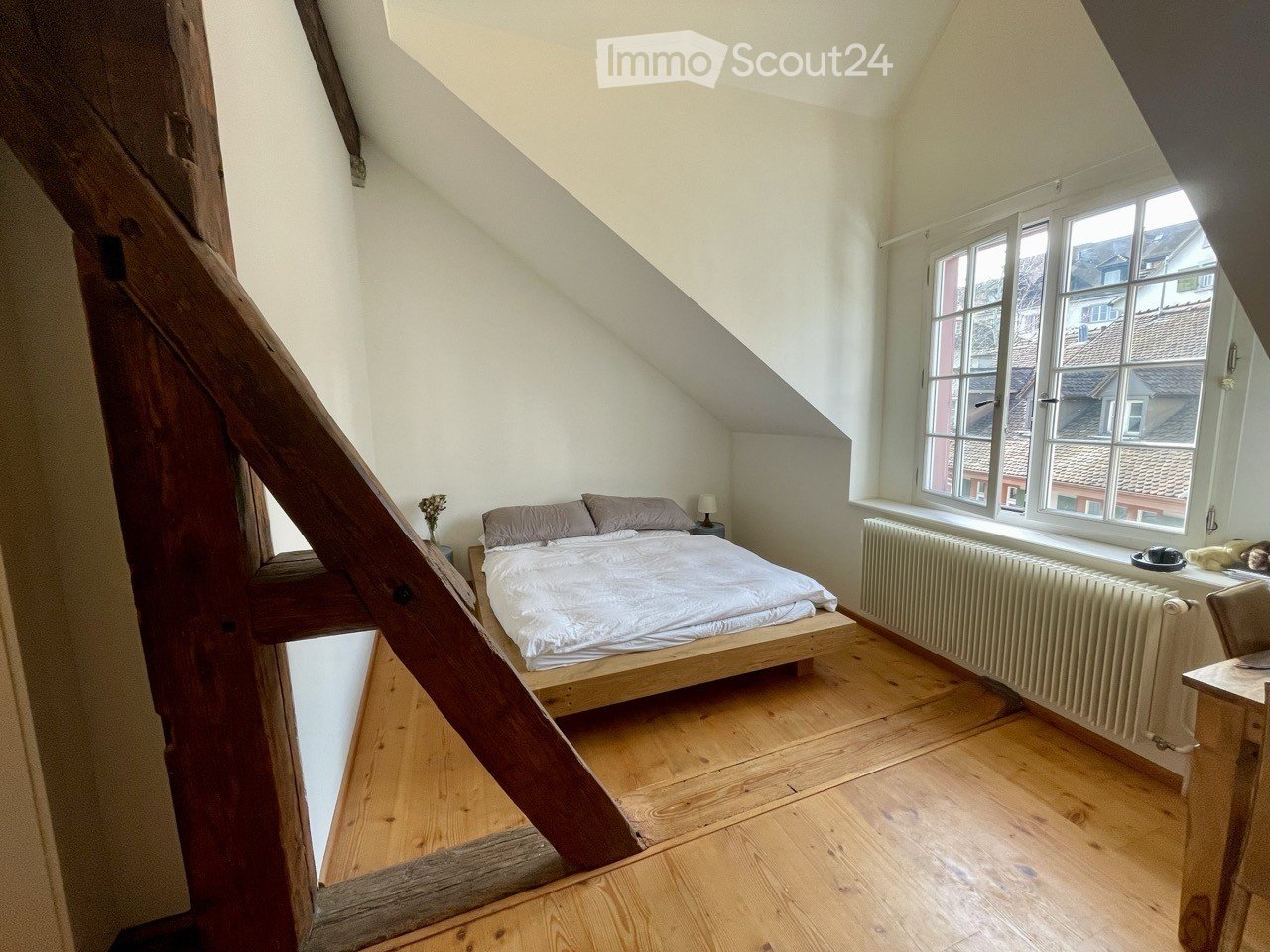 Cozy attic bedroom with wooden floors, slanted ceilings, large windows, a bed with pillows and a duvet, a nightstand with a lamp, a radiator, and a small table with a chair.