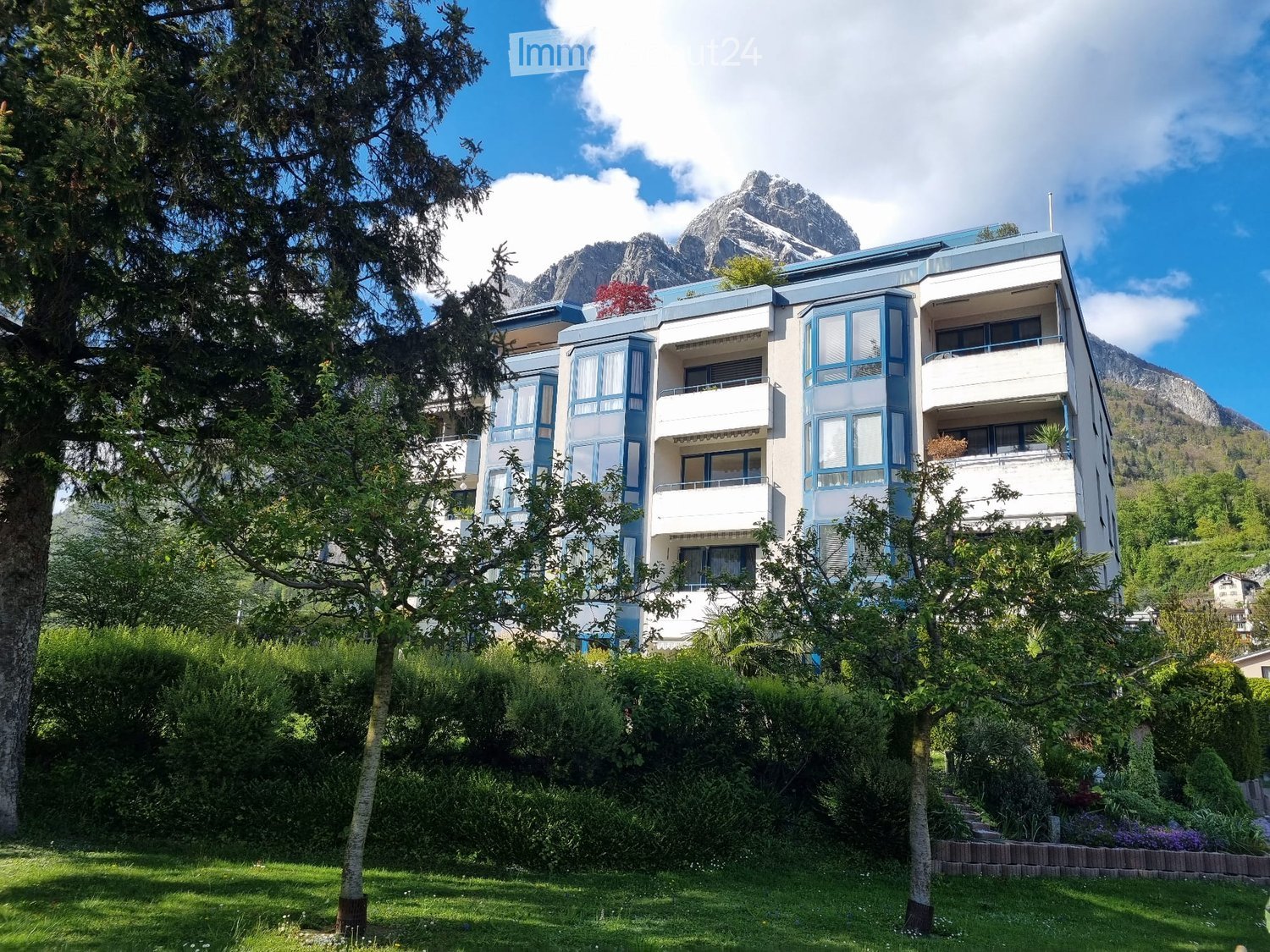 Modern multi-story building with white and blue facade, large windows, and balconies, surrounded by lush greenery and trees, with a mountain view in the background.
