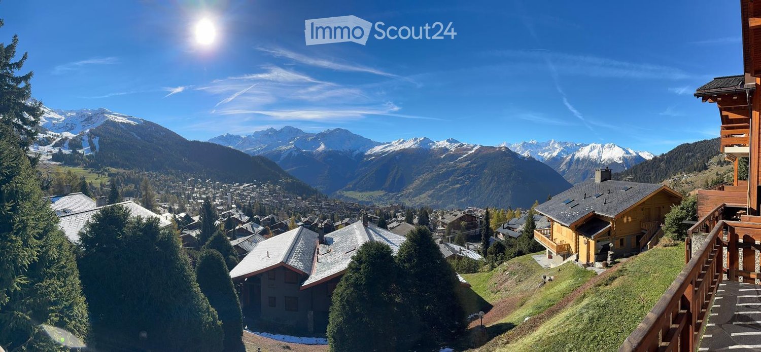Panoramic view of a mountain village with snow-capped peaks, wooden chalets, and a clear blue sky.
