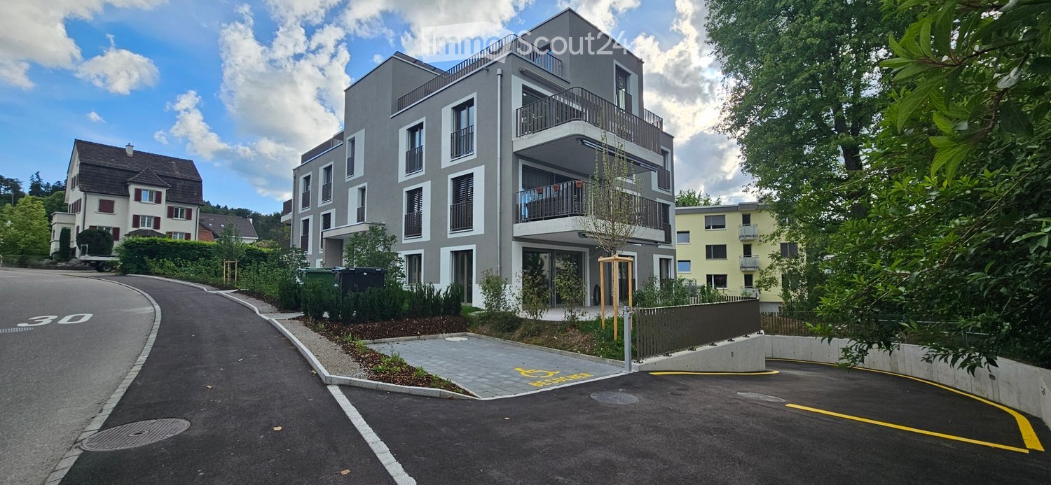 Modern multi-story apartment building with balconies, surrounded by greenery and a parking area.
