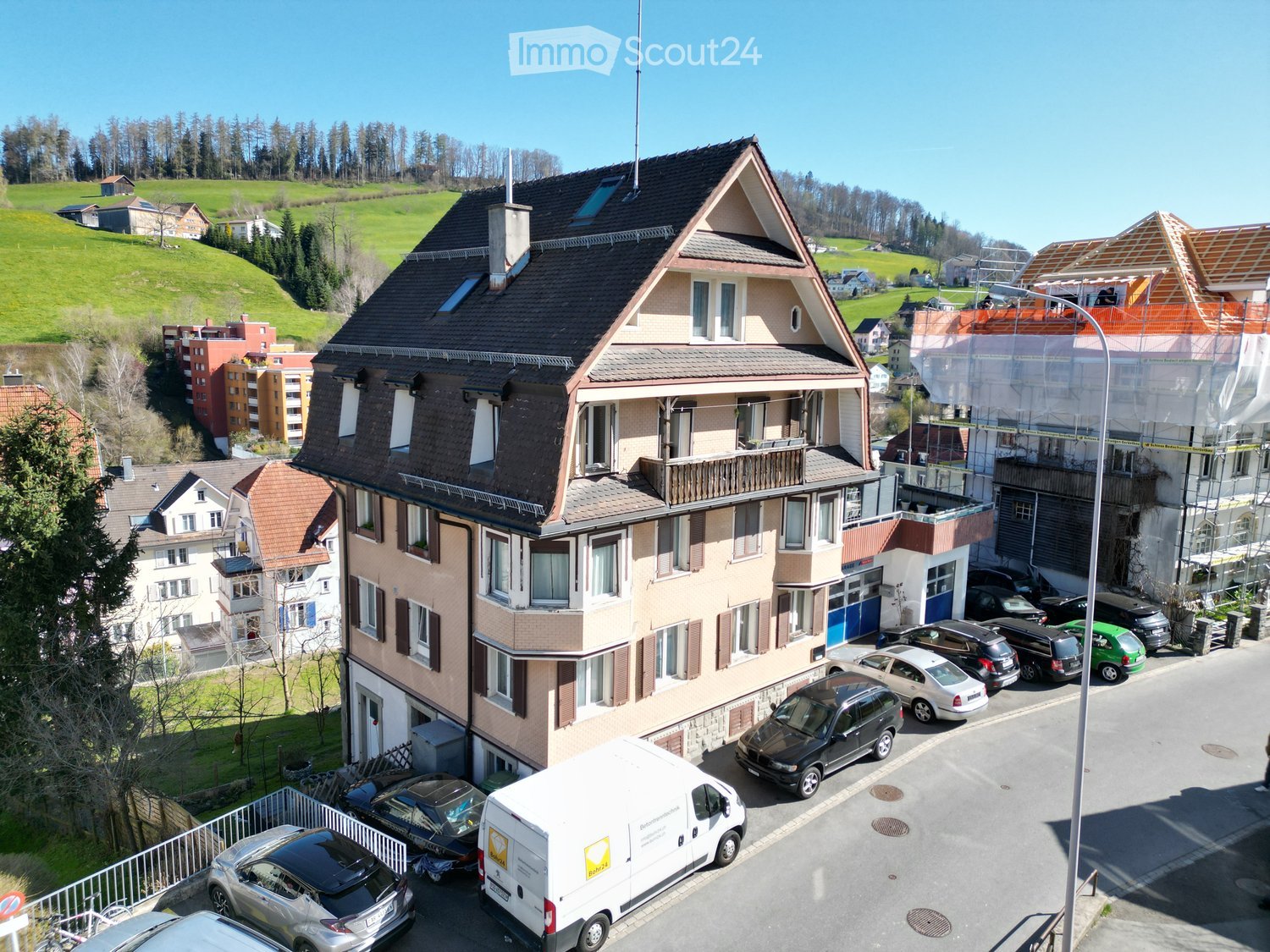 A multi-story residential building with a pitched roof, multiple windows, and a balcony on the upper floor. The building is surrounded by parked cars and other residential structures in a suburban area with green hills in the background.