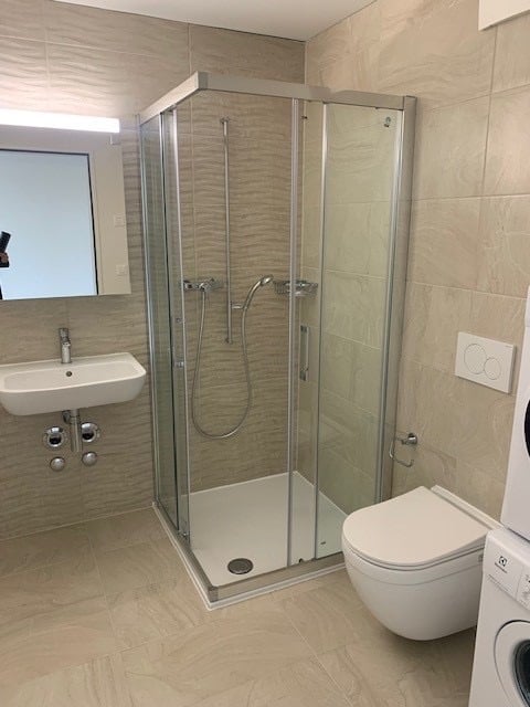 Modern bathroom with a glass-enclosed shower stall, a white toilet, a sink with a mirror, and a washing machine visible in the corner.