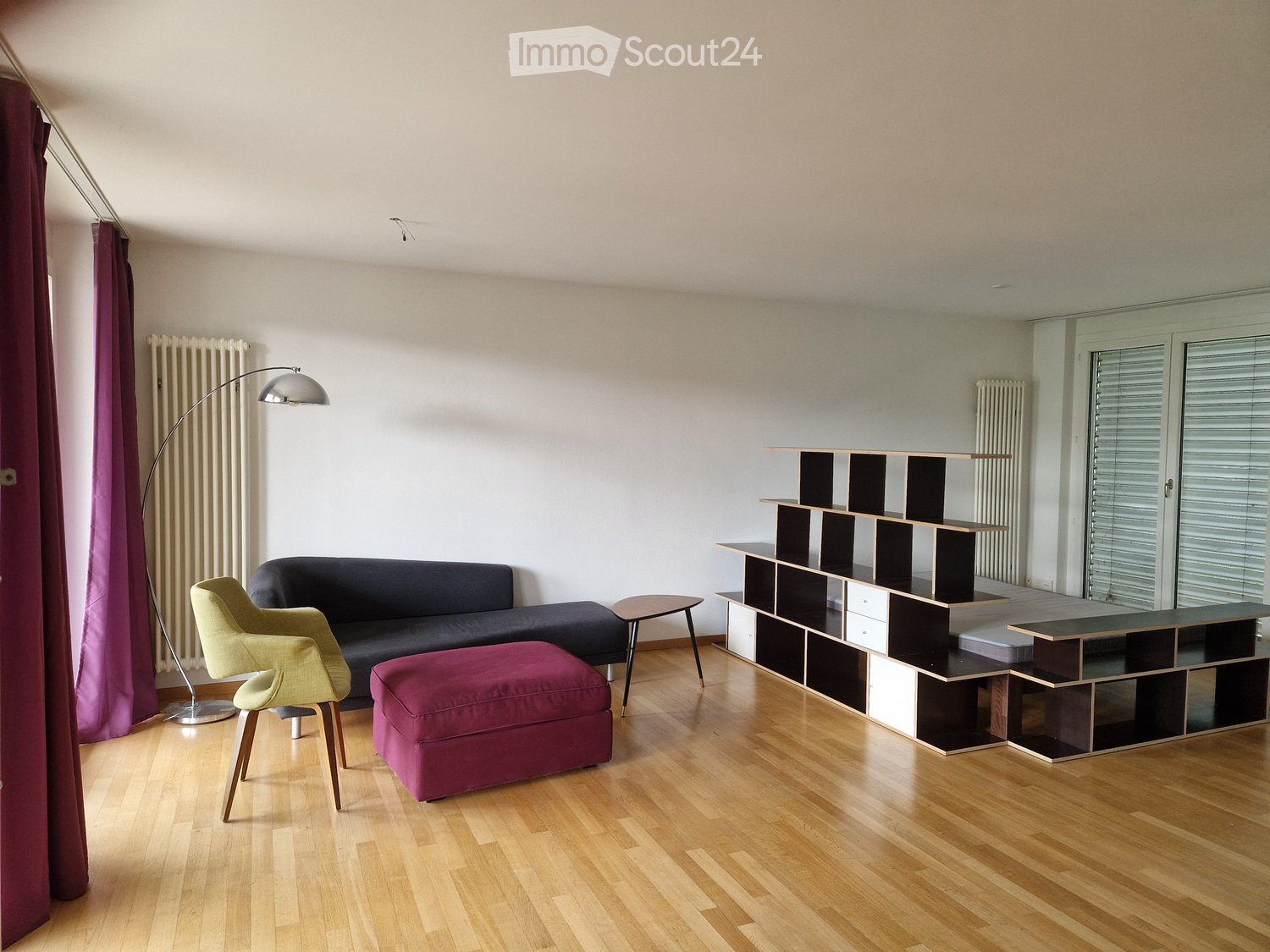 Modern living room with light wooden flooring, a dark grey sofa, a yellow armchair, a purple ottoman, a small wooden side table, and a modular shelving unit.