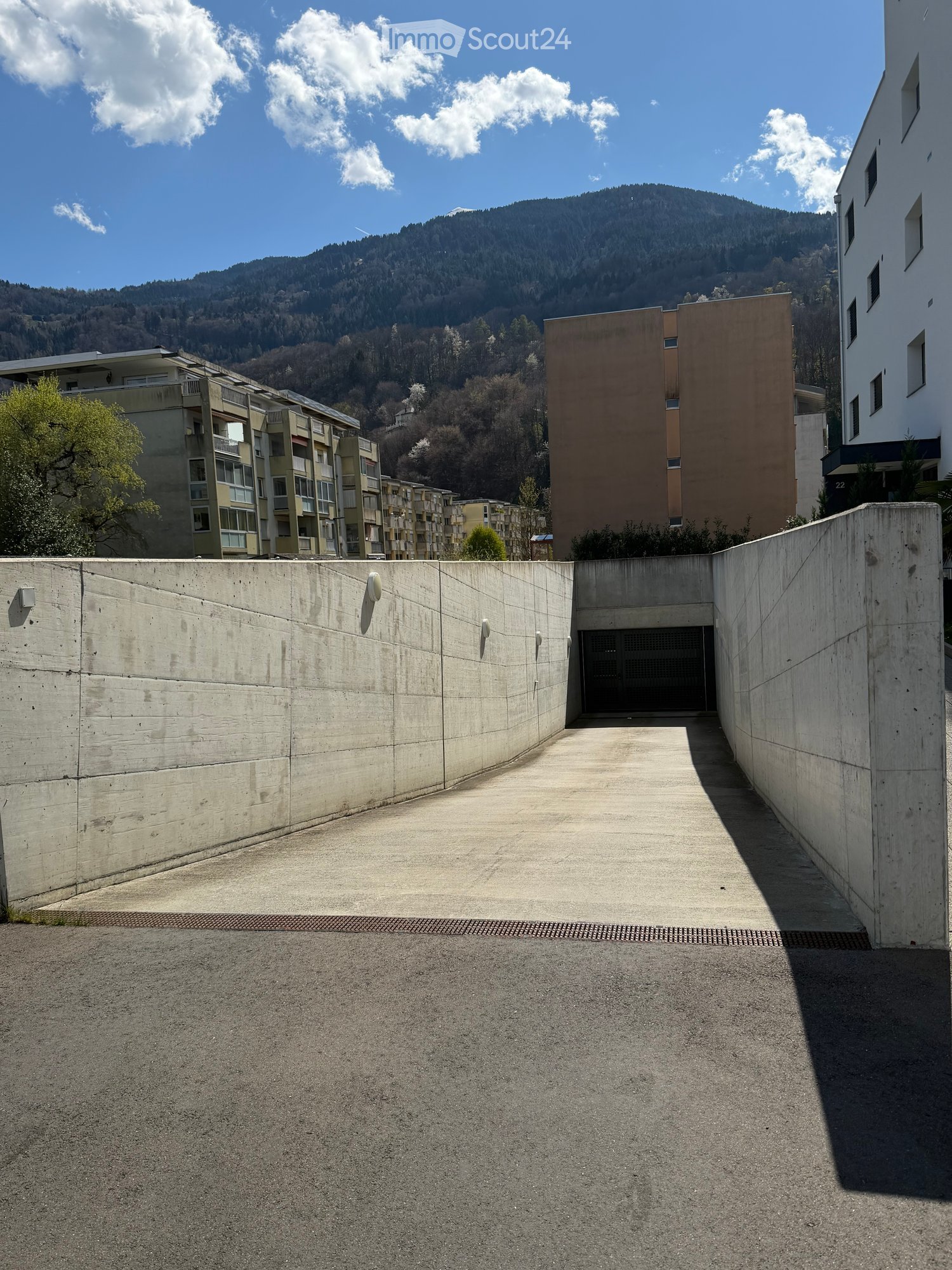 A modern residential complex with a raised ground floor entrance, surrounded by concrete walls and a scenic mountain backdrop.
