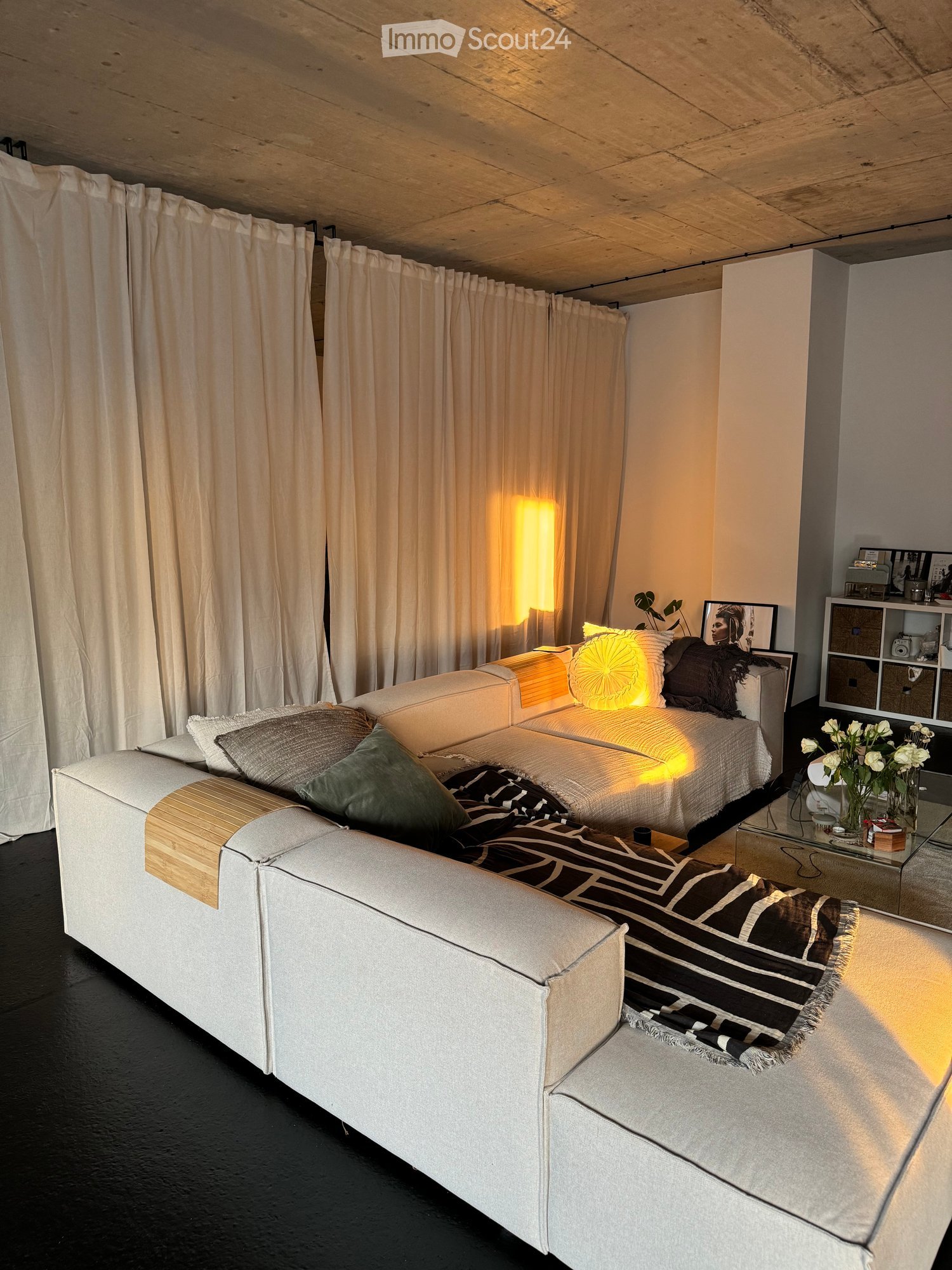 Modern living room with a white sectional sofa, throw pillows, a coffee table with a vase of white flowers, a table lamp, and a shelf with decorative items.
