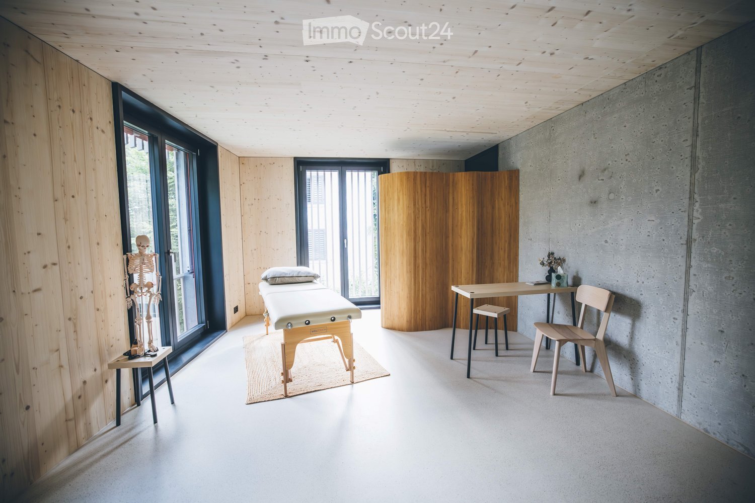 Modern, minimalist room with light wood and concrete finishes, featuring a massage table, skeleton model, small table, and chairs.
