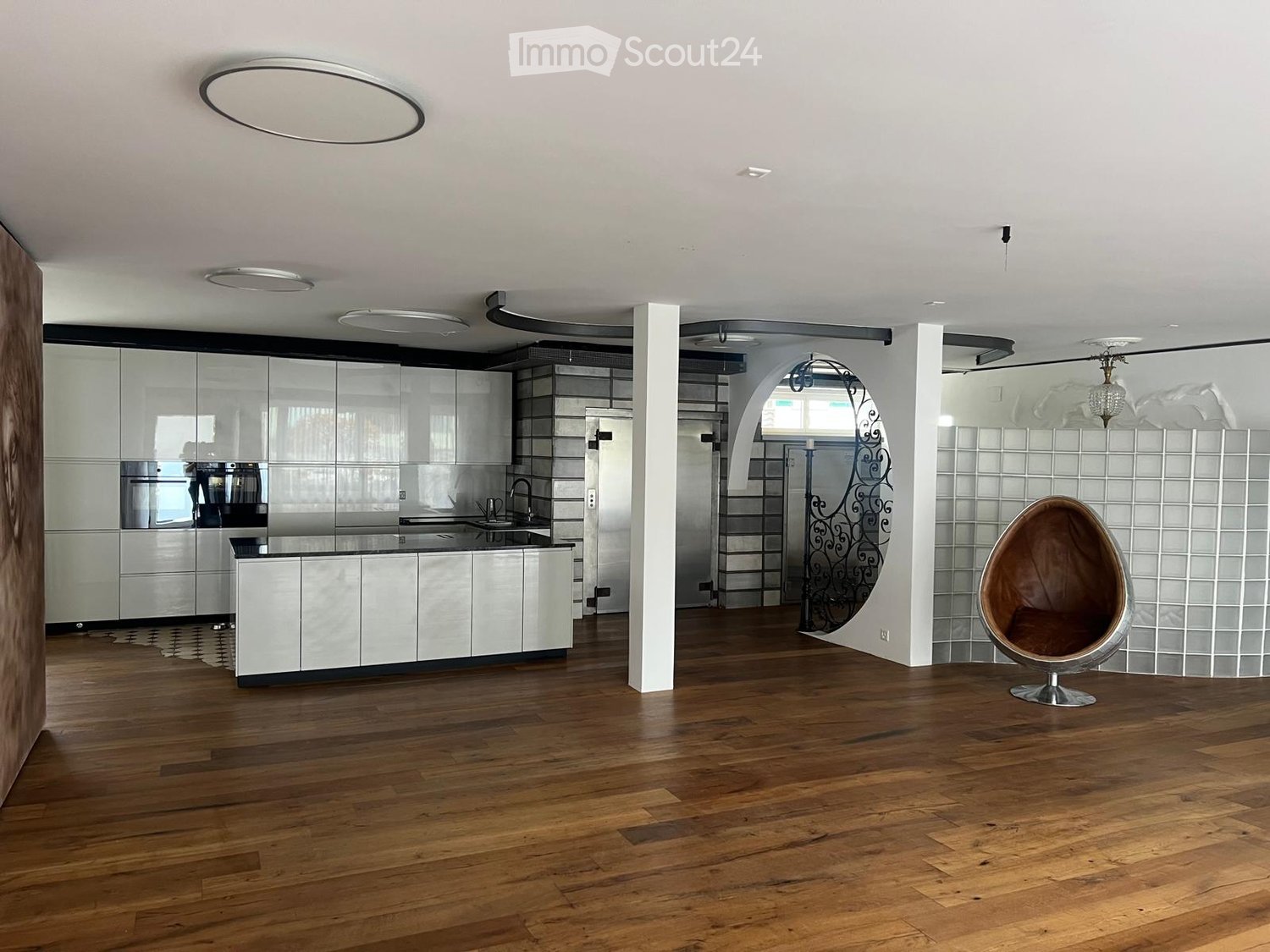 Modern, open-plan space with a kitchen area and a living area. Features include sleek white cabinets, a stainless steel sink, a round egg-shaped chair, and a polished wooden floor.