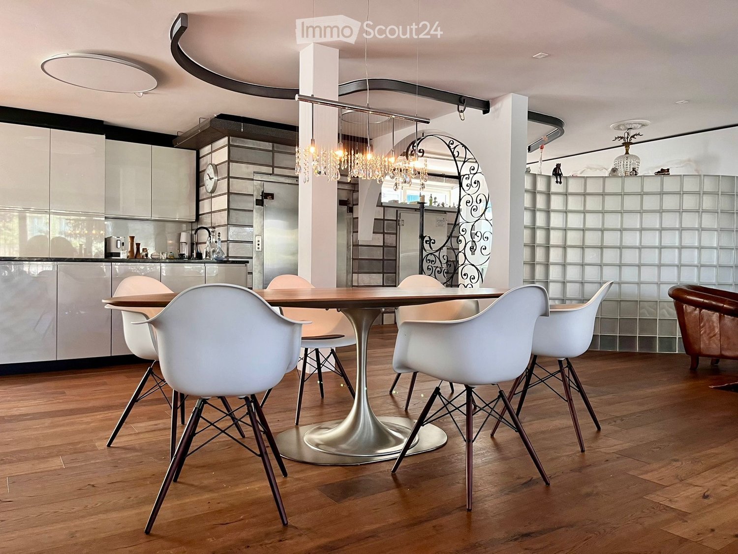 Modern dining area with a round wooden table, white chairs, and a sleek kitchen area in the background.