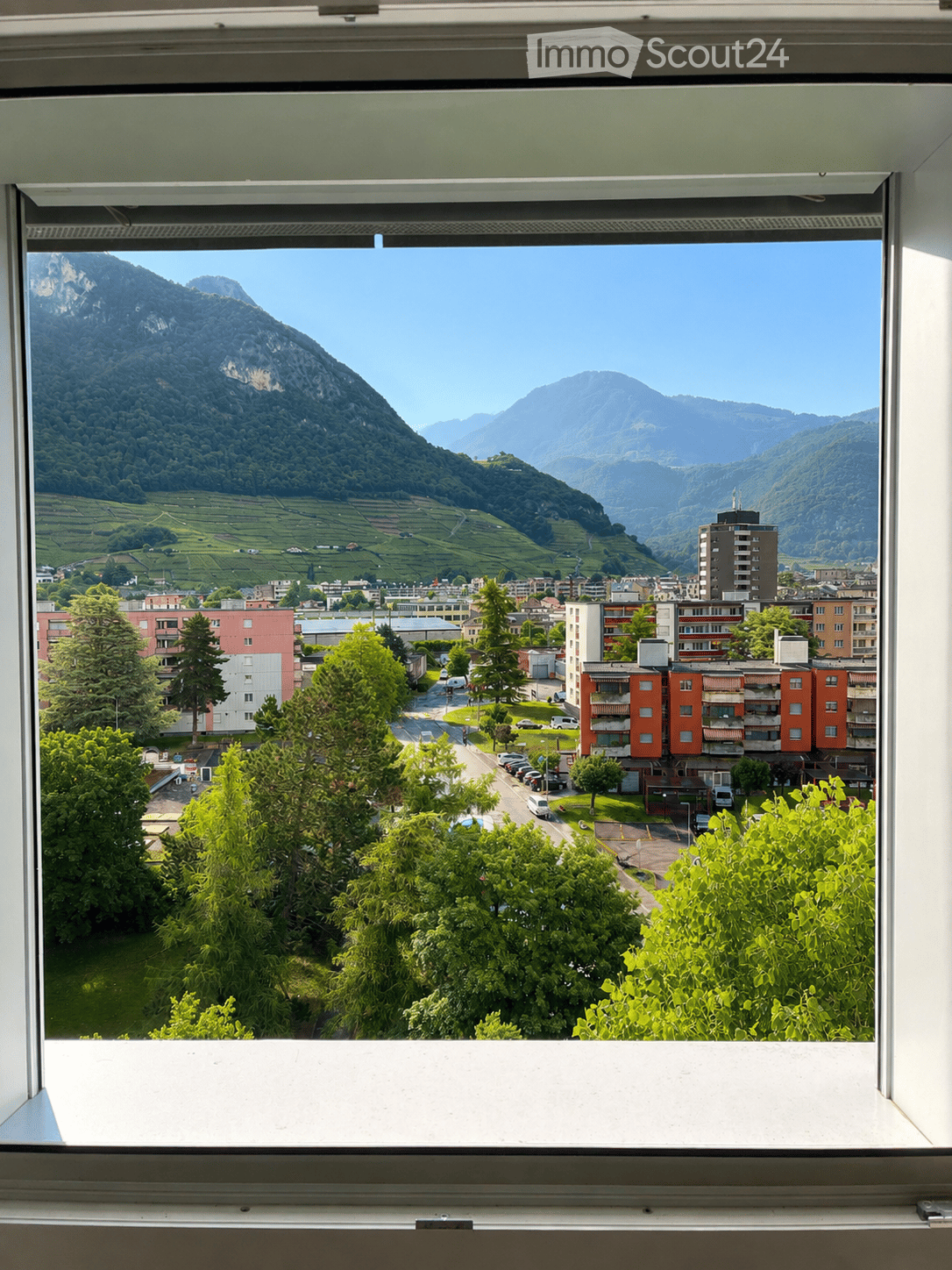 View from a window showing a town with buildings, trees, and distant mountains.
