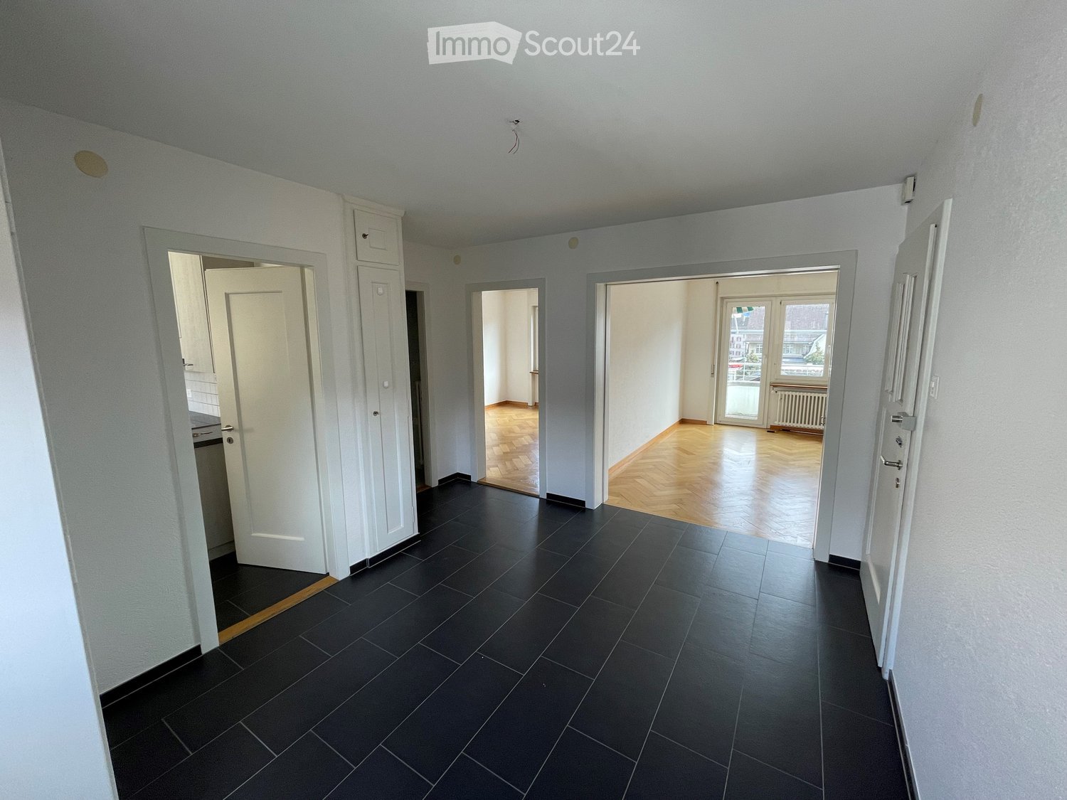 Empty hallway with dark tiled floor and white walls, leading to multiple rooms with wooden flooring and large windows.