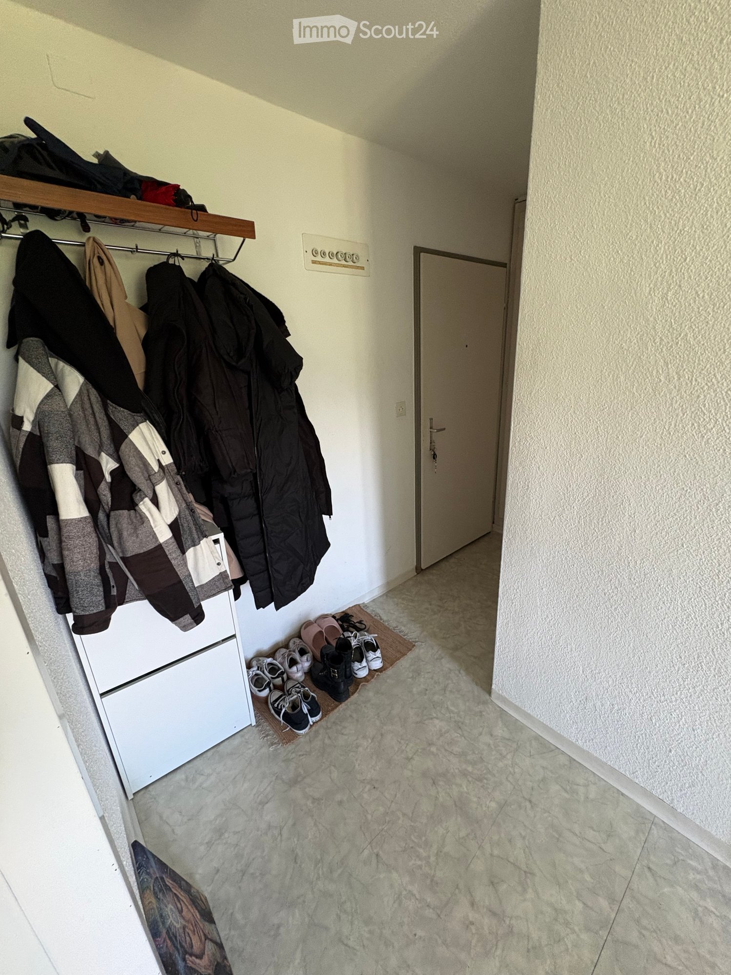 A narrow hallway with a coat rack holding jackets, a shelf above it, and a row of shoes placed on a small mat near the door.