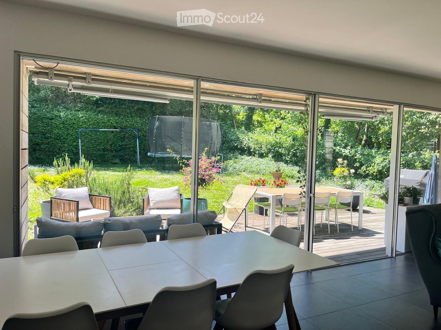 Bright room with large sliding glass doors opening to a garden, featuring a dining table with chairs, a sofa with pillows, and an ottoman.
