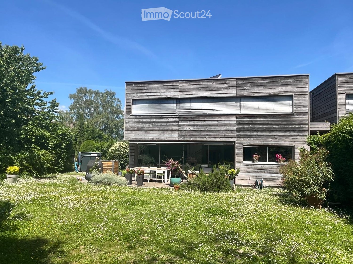 Modern two-story wooden house with large windows and a spacious garden area with a patio and outdoor furniture.