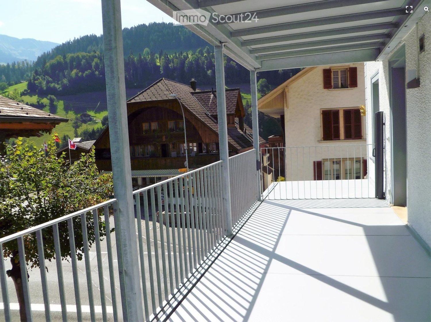 Balcony with white metal railing, overlooking a scenic valley with houses and mountains in the background.