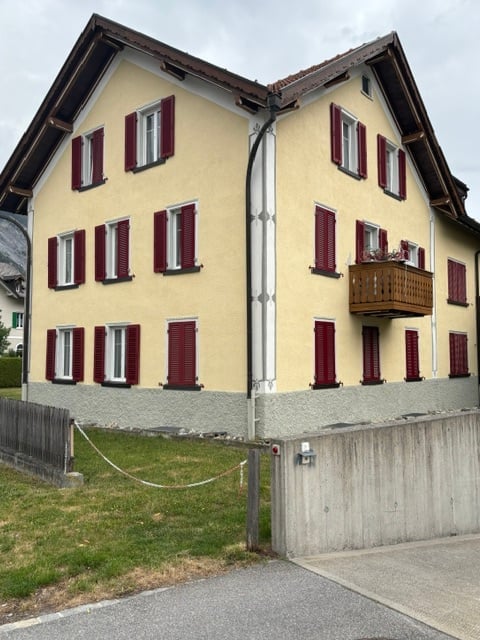Two-story house with yellow walls, red shutters, and a wooden balcony on the second floor.