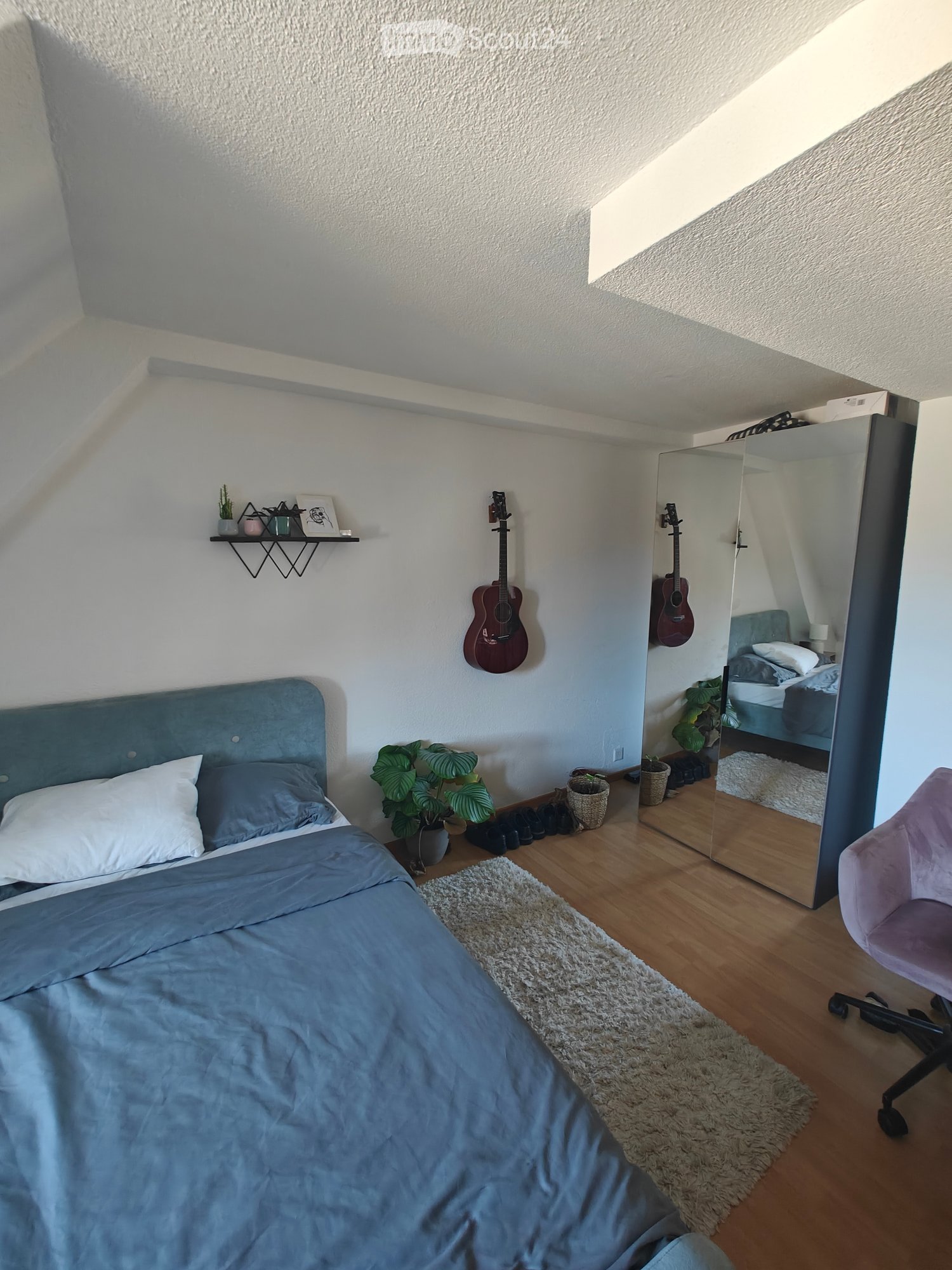 Cozy bedroom with a blue bed, wooden floor, large mirror, guitars, and a small shelf with decorative items.