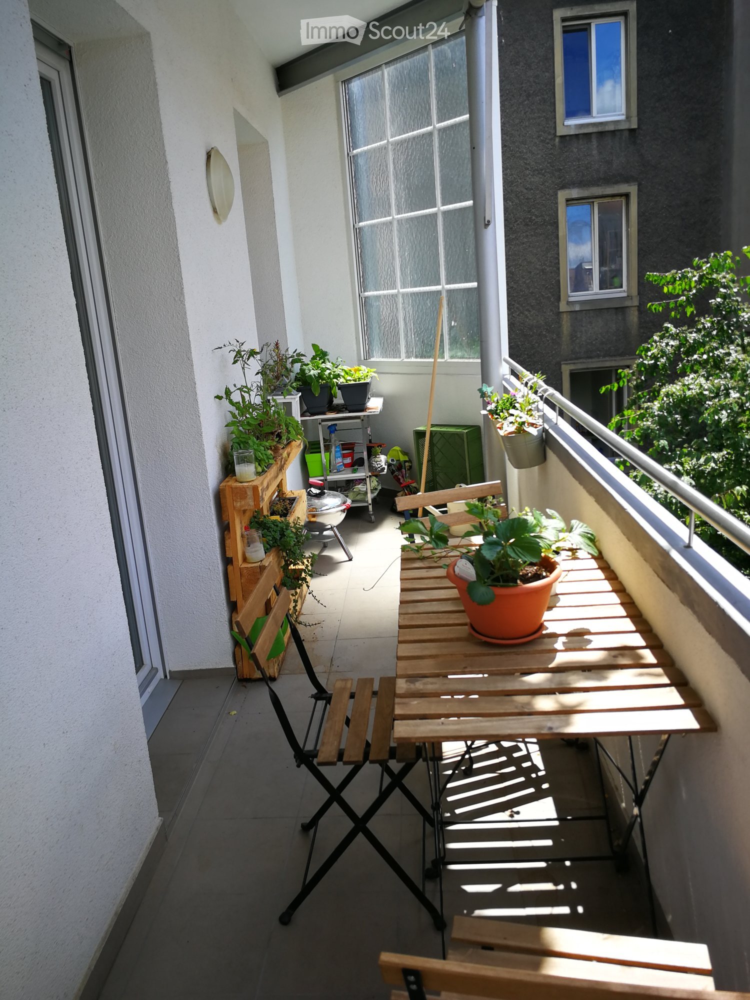 Balcony with wooden table, folding chair, potted plants, and railing overlooking greenery.