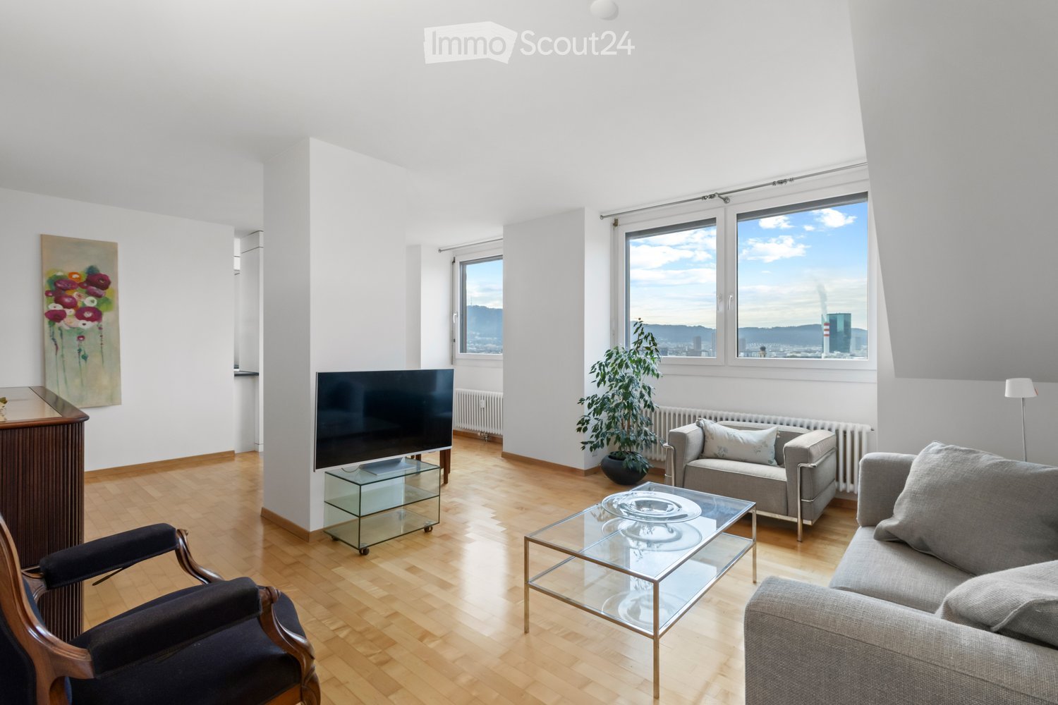 Living Room. Spacious living room with wooden floors, large windows, and a view of the city. Features a couch, chair, coffee table, TV, and a potted plant. A painting adorns the wall, and a lamp is placed on the side.