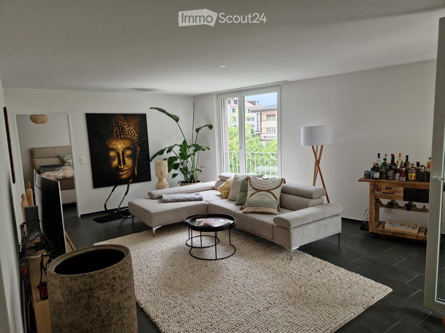 Modern living room with a grey sectional sofa, coffee table, floor lamp, large mirror, artwork, and a bar cart with bottles.
