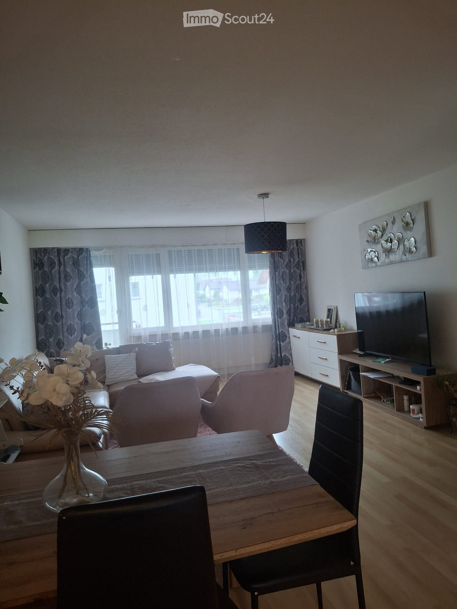 Living room with a sofa, chairs, a wooden table, a vase with flowers, a television, and a cabinet. The room features large windows with curtains and a decorative wall piece.