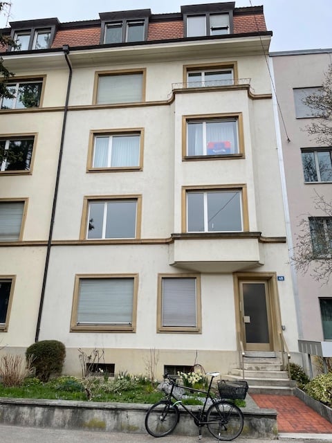 Three-story apartment building with white exterior, multiple windows, two bicycles parked in front, a small garden area with plants in front of the building.