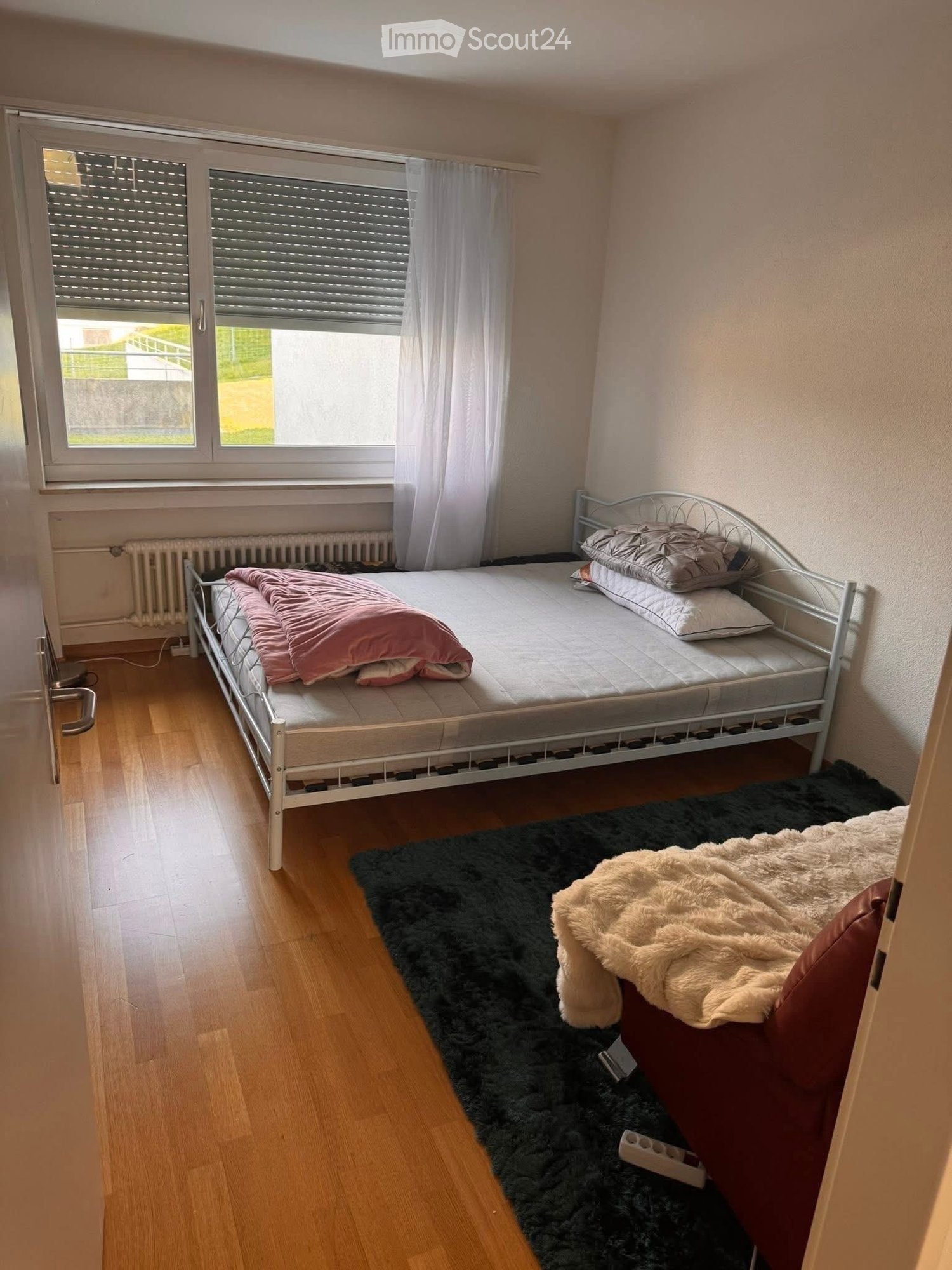 Cozy bedroom with wooden flooring, a single bed with pillows and a blanket, a fluffy rug, and large windows with blinds and curtains.